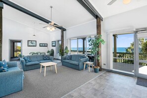 Ein geräumiges Wohnzimmer mit zwei Sofas, einem Couchtisch und einem Deckenventilator. Große Fenster bieten Blick auf das Meer.