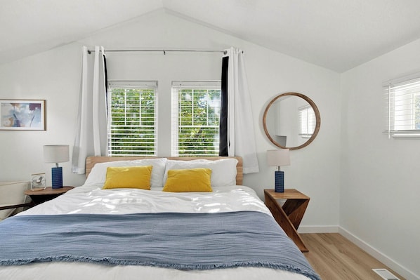 A bedroom with a bed, two bedside tables, a mirror, and a window with blinds.