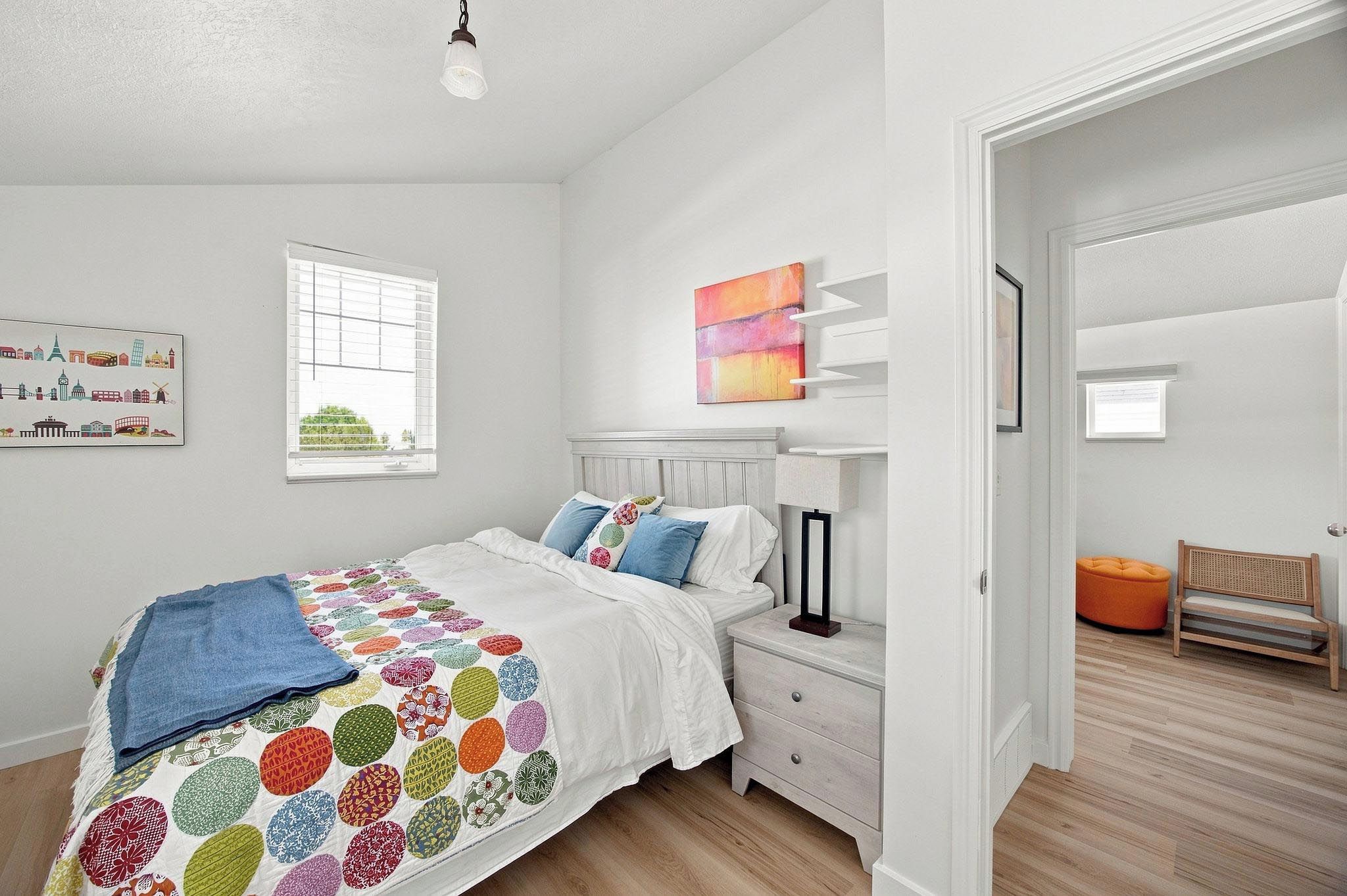 Un dormitorio con cama, mesitas de noche, una ventana y una puerta que da a otra habitación.