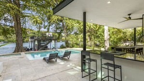 Un espace piscine extérieur moderne, doté de chaises longues et d’un bar, offrant une vue sur un lac et entouré d’arbres.