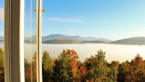 Blick durch ein Fenster auf eine Berglandschaft mit Herbstlaub und einer Nebelschicht am Fuße der Berge.