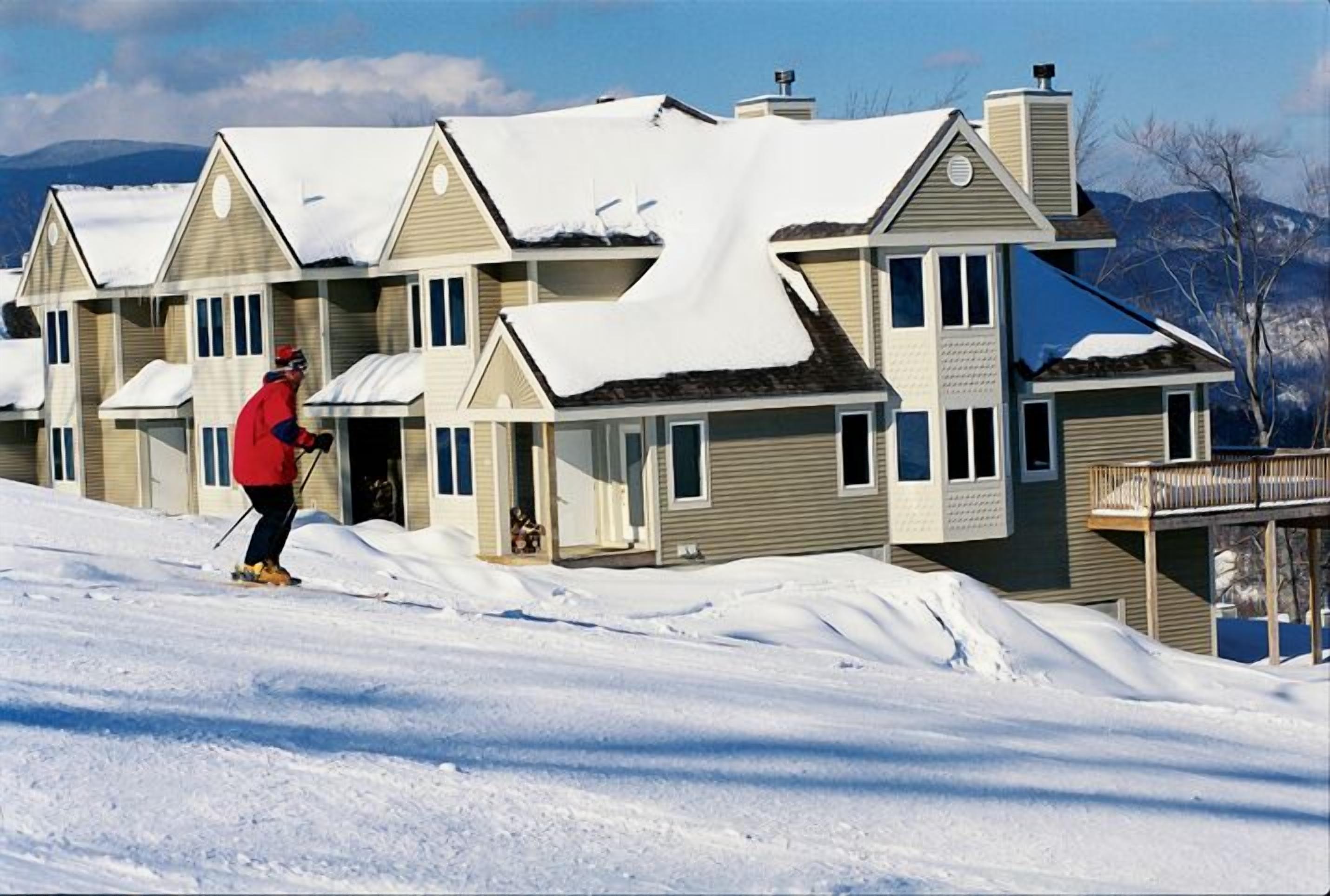 A row of two-story houses with snow-covered roofs, a person skiing in the foreground, and mountains in the background.