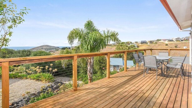 A wooden deck with a table and chairs, overlooking a coastal landscape with houses and hills.