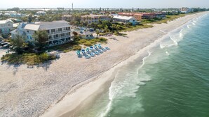 Ein Strandhotel mit mehreren Gebäuden, einem Sandstrand und einer Reihe blauer Liegen und Sonnenschirme.