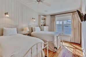 Een slaapkamer met twee bedden, een nachtkastje, een dressoir, een raam met gordijnen en een plafondventilator.
