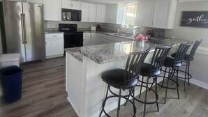 A modern kitchen with white cabinetry, a stainless steel refrigerator, a black microwave, and a granite countertop island with four black bar stools. - COMPLETELY REMODLED!! Gorgeous 5 BR/5.5 BA in NO WAKE cove, Dock w/12'x30' slip (Sunrise Beach)