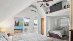 Une chambre avec un grand lit, un ventilateur de plafond, un climatiseur, un téléviseur posé sur une étagère et un tableau représentant des voiliers.