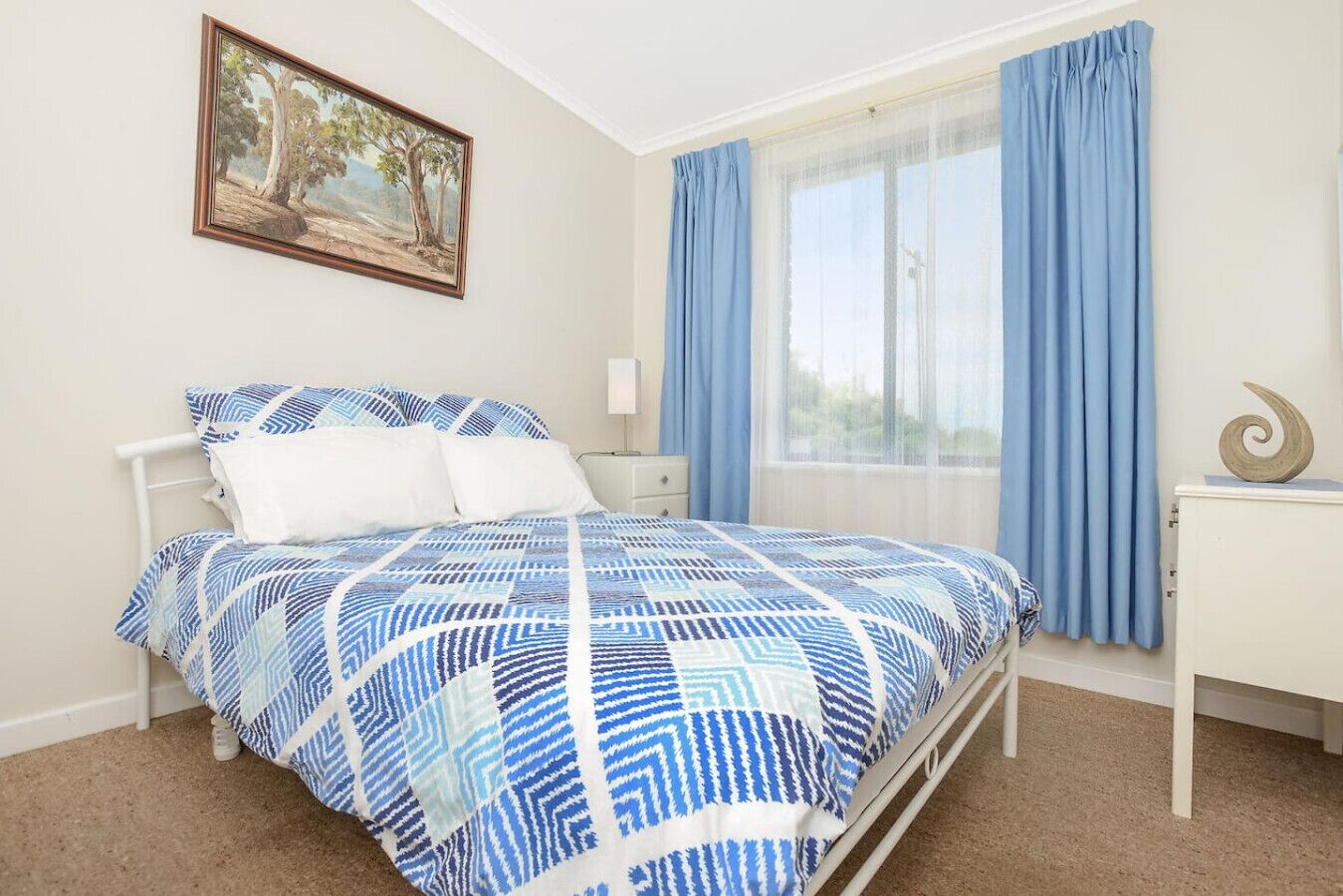 Un dormitorio con cama, mesita de noche, lámpara, ventana con cortinas y un cuadro en la pared.