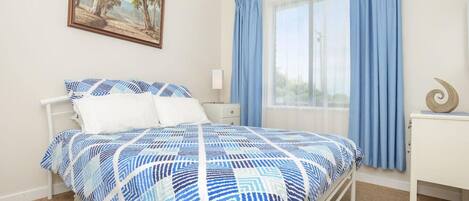 Un dormitorio con cama, mesita de noche, lámpara, ventana con cortinas y un cuadro en la pared.