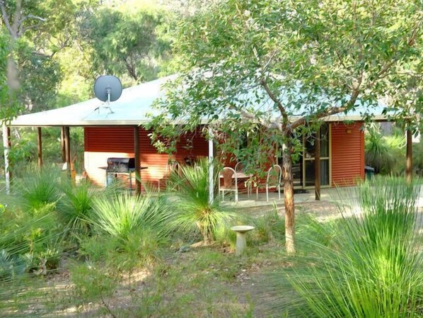 A rustic cabin with a satellite dish, surrounded by lush greenery and a small patio area.