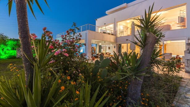 Ein modernes, zweistöckiges Haus mit Balkon, umgeben von üppiger Vegetation und blühenden Pflanzen.