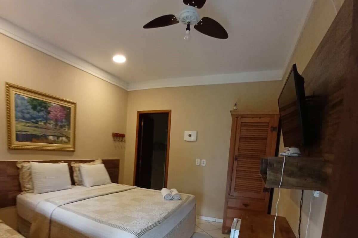 Quarto com cama, ventilador de teto, pintura, guarda-roupa de madeira e televisão sobre um suporte de madeira.