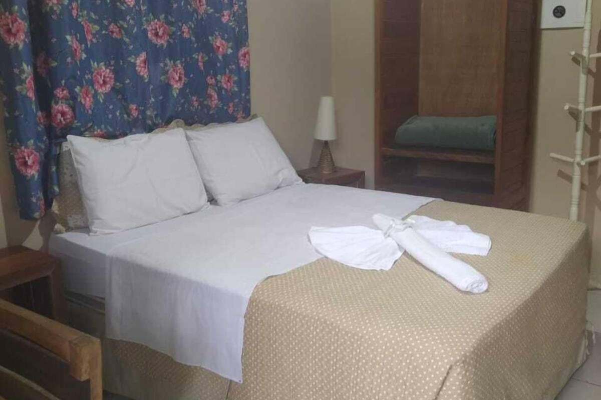 Uma cama de solteiro com lençóis brancos, uma mesa de cabeceira de madeira com abajur, um guarda-roupa de madeira e uma cortina florida.