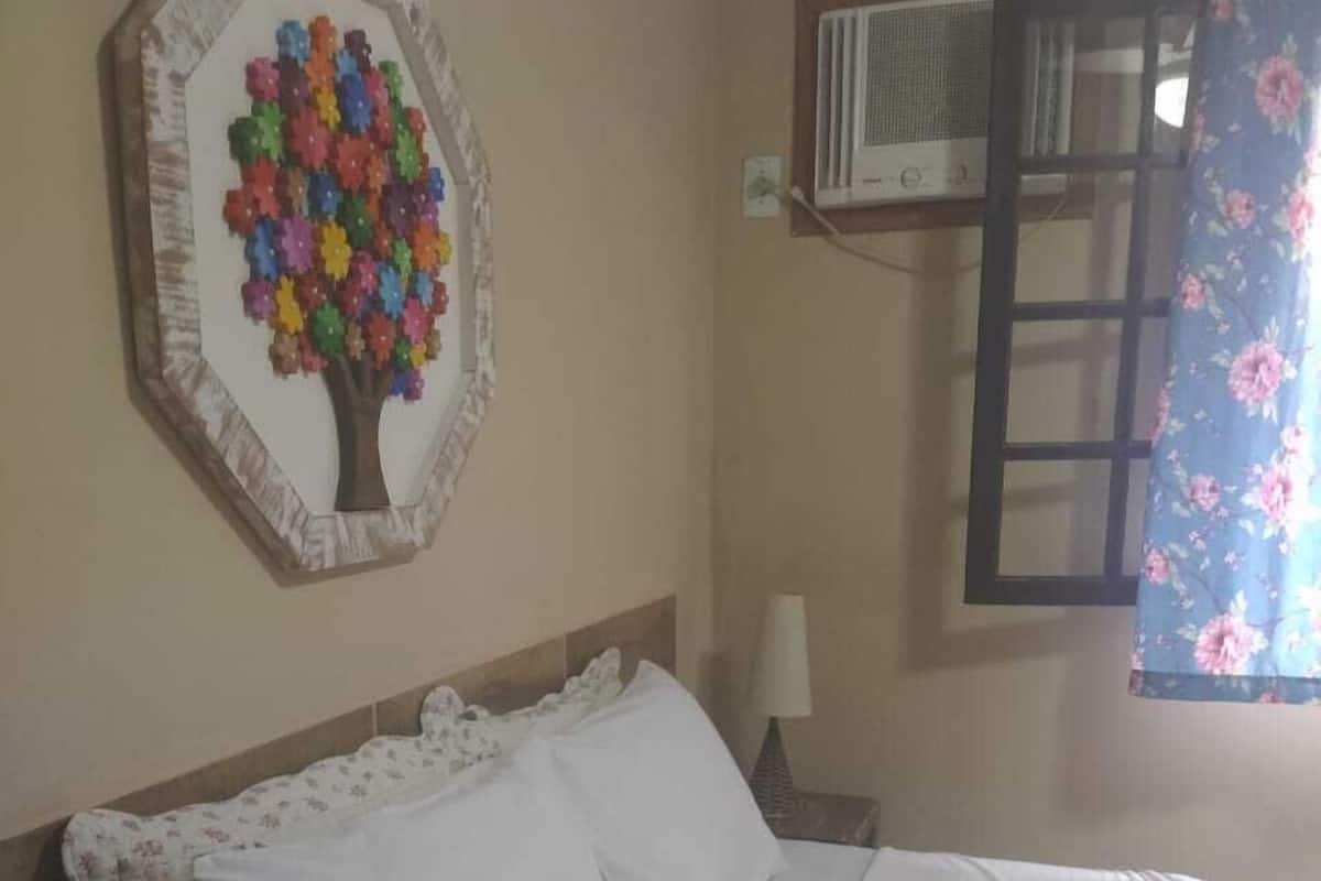 Quarto com cama, decoração floral na parede, janela com cortina florida e ar condicionado.