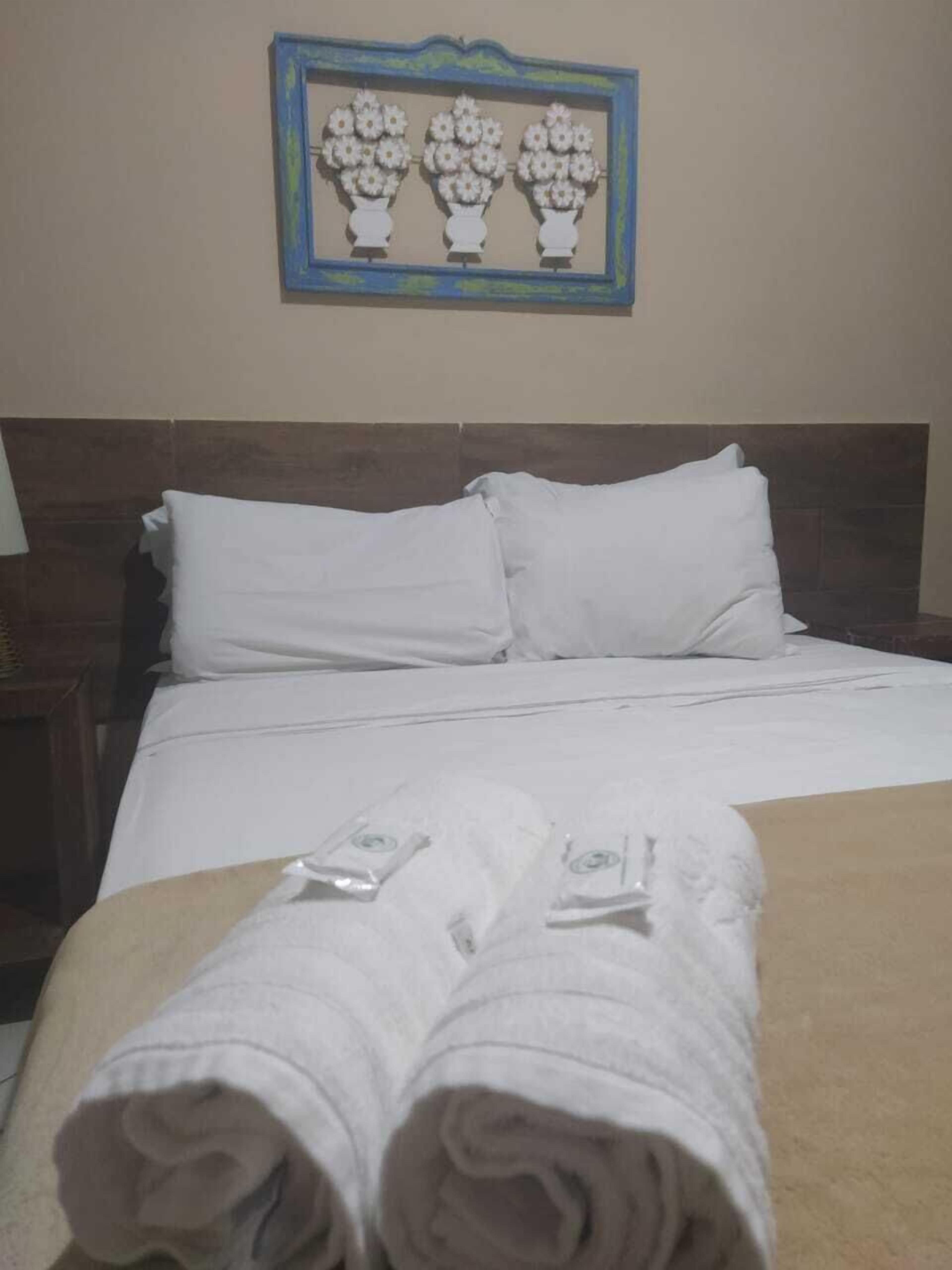 A neatly made bed with white linens and towels, a wooden headboard, and a framed picture of flowers on the wall.