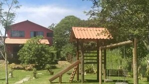 Un espace extérieur verdoyant avec une structure de jeux en bois, un bâtiment rouge en arrière-plan et un ciel bleu dégagé.