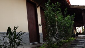 Un chemin mène à un bâtiment doté d’un toit en tuiles et d’une porte. On y trouve des plantes en pot et un petit espace jardin agrémenté de pas japonais.