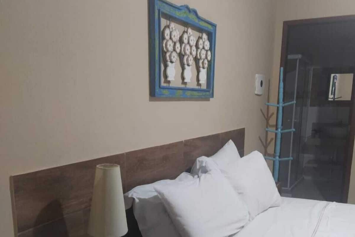 Cama bem arrumada com roupa de cama branca, mesa de cabeceira de madeira com abajur e uma obra de arte na parede.