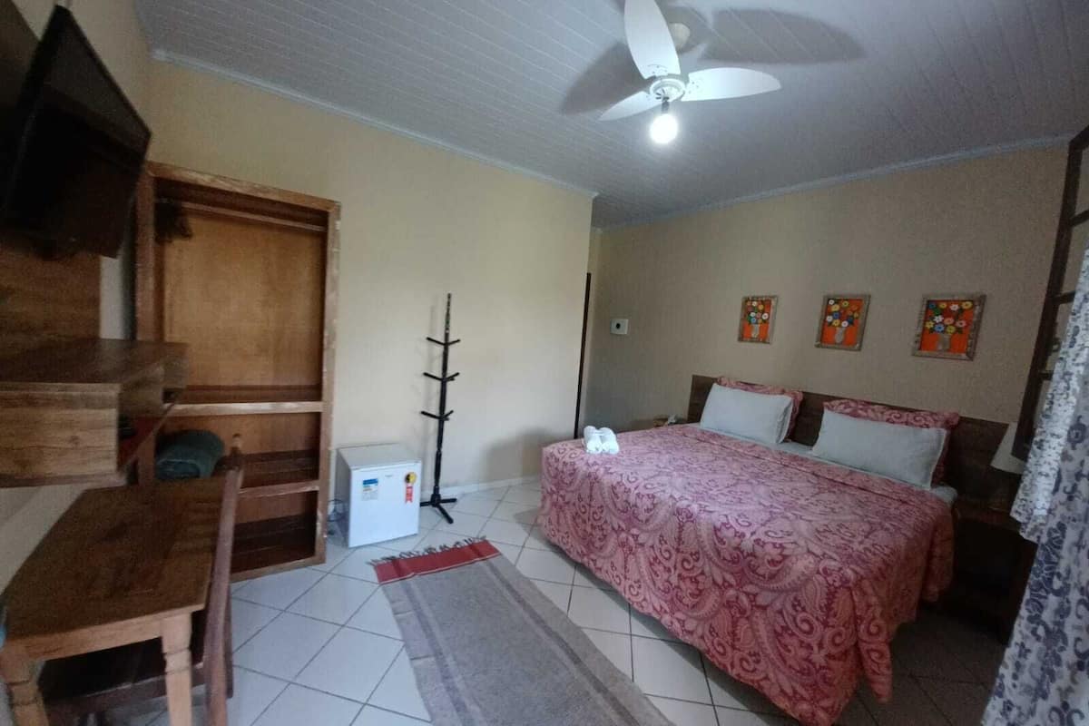 Quarto com cama, escrivaninha, cadeira, ventoinha no teto e radiador.