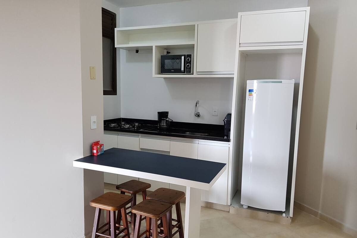 A compact kitchen with a black countertop, white cabinets, a microwave, a refrigerator, and three wooden stools.