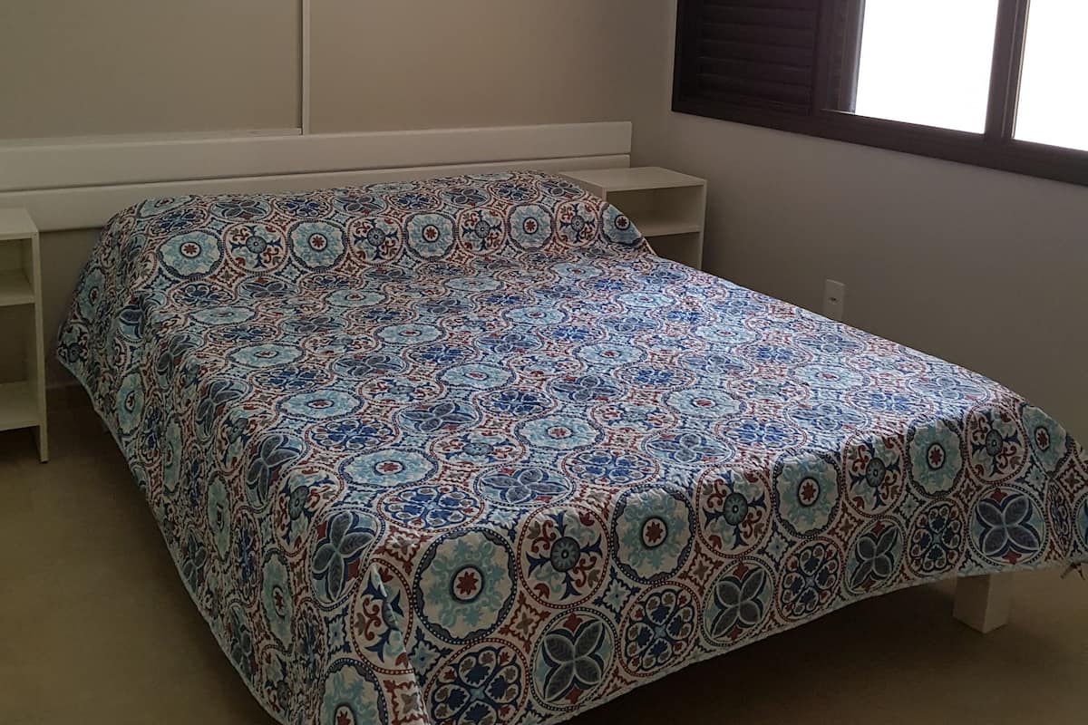 A bedroom with a bed, a window with blinds, an air conditioner, and a framed picture on the wall.