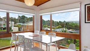 Een eetgedeelte met een glazen tafel en witte stoelen, grote ramen met uitzicht op huizen en begroeiing.