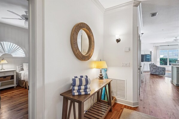 A well-lit hallway with a wooden console table, a mirror, and a lamp. A bed with a blue and white striped blanket is visible in the background.