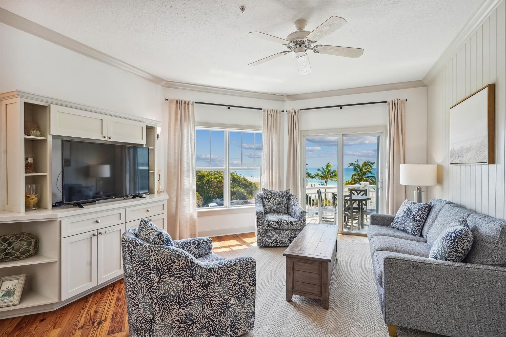 A living room with a television, two patterned armchairs, a sofa, and a coffee table. There is a ceiling fan, large windows with curtains, and a view of the outdoors.