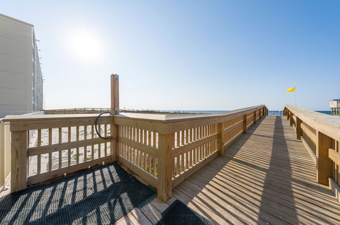 Ein Holzsteg führt zu einem Strand, mit blauem Himmel und einer Wimpel im Hintergrund.