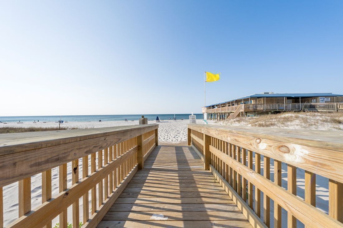 Ein hölzerner Steg führt zu einem Strand mit einer gelben Fahne, einem Gebäude mit Balkon und einem wolkenlosen Himmel.