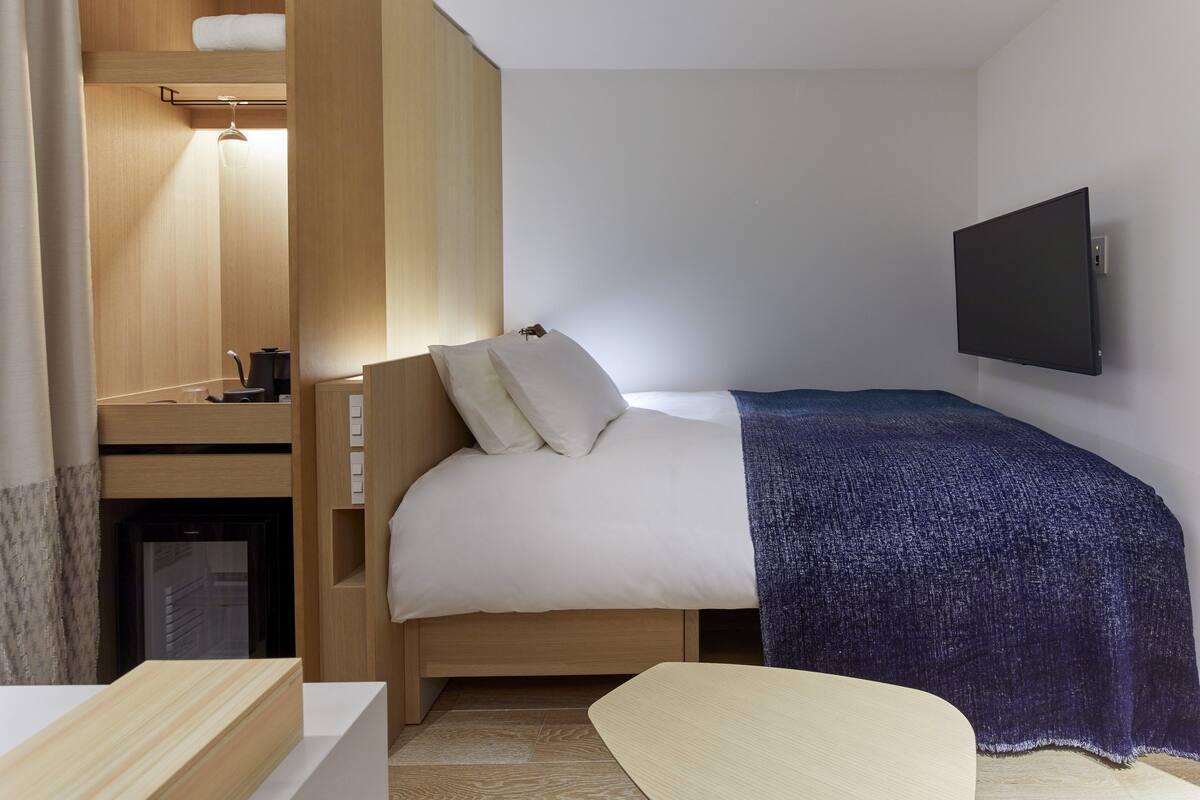 A compact hotel room with a single bed, a small desk with a chair, a television mounted on the wall, and a compact kitchenette area.
