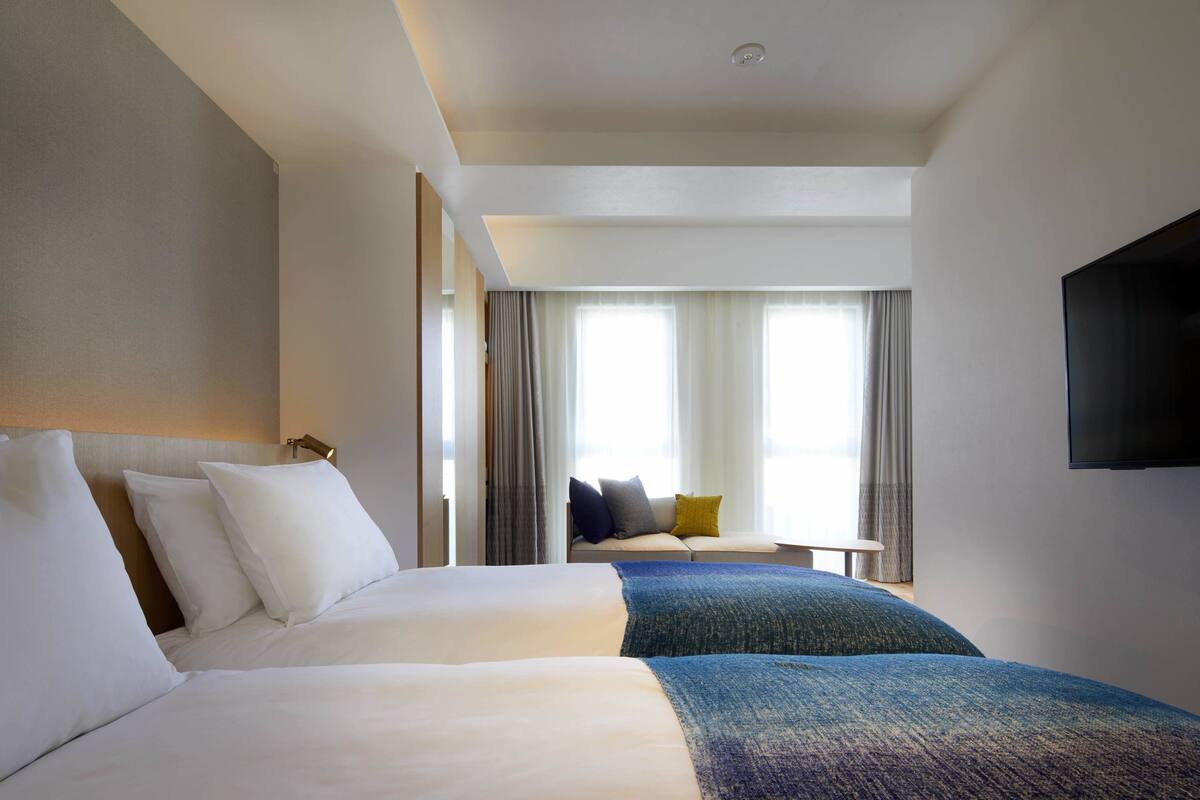 A hotel room with a large bed, a smaller bed, a TV mounted on the wall, and a window with curtains.