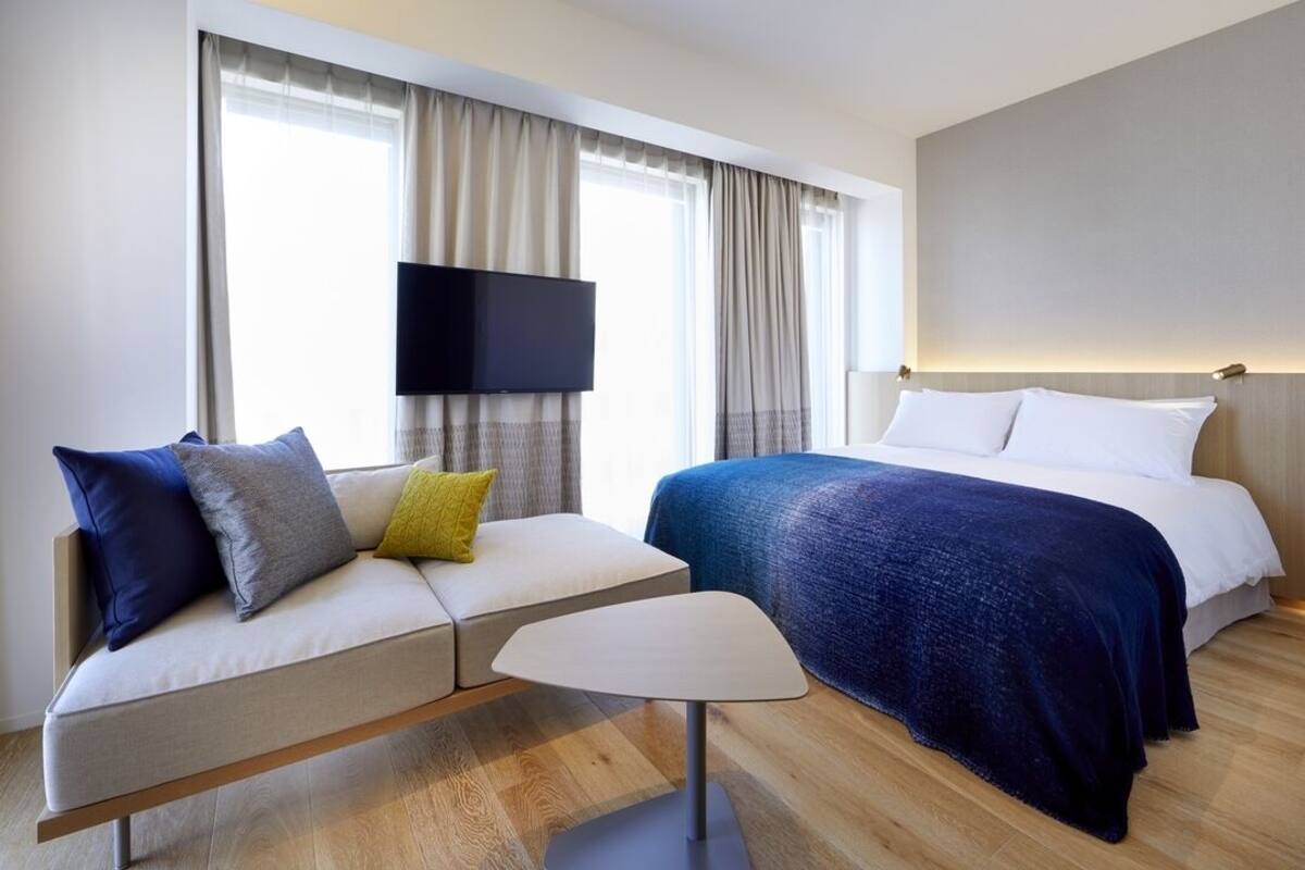 A modern hotel room with a sofa, a small table, a bed, and a TV mounted on the wall.