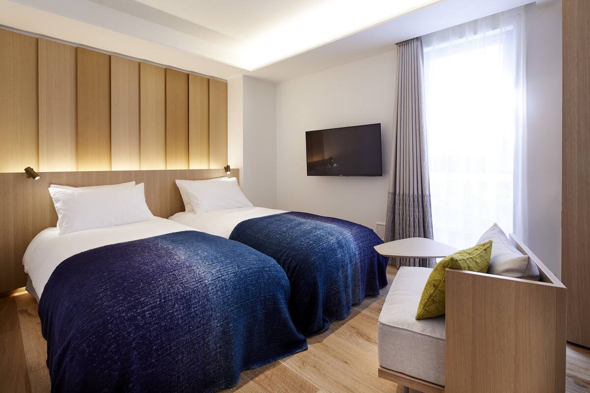 A hotel room with two beds, a TV, a small table, and a window with curtains.