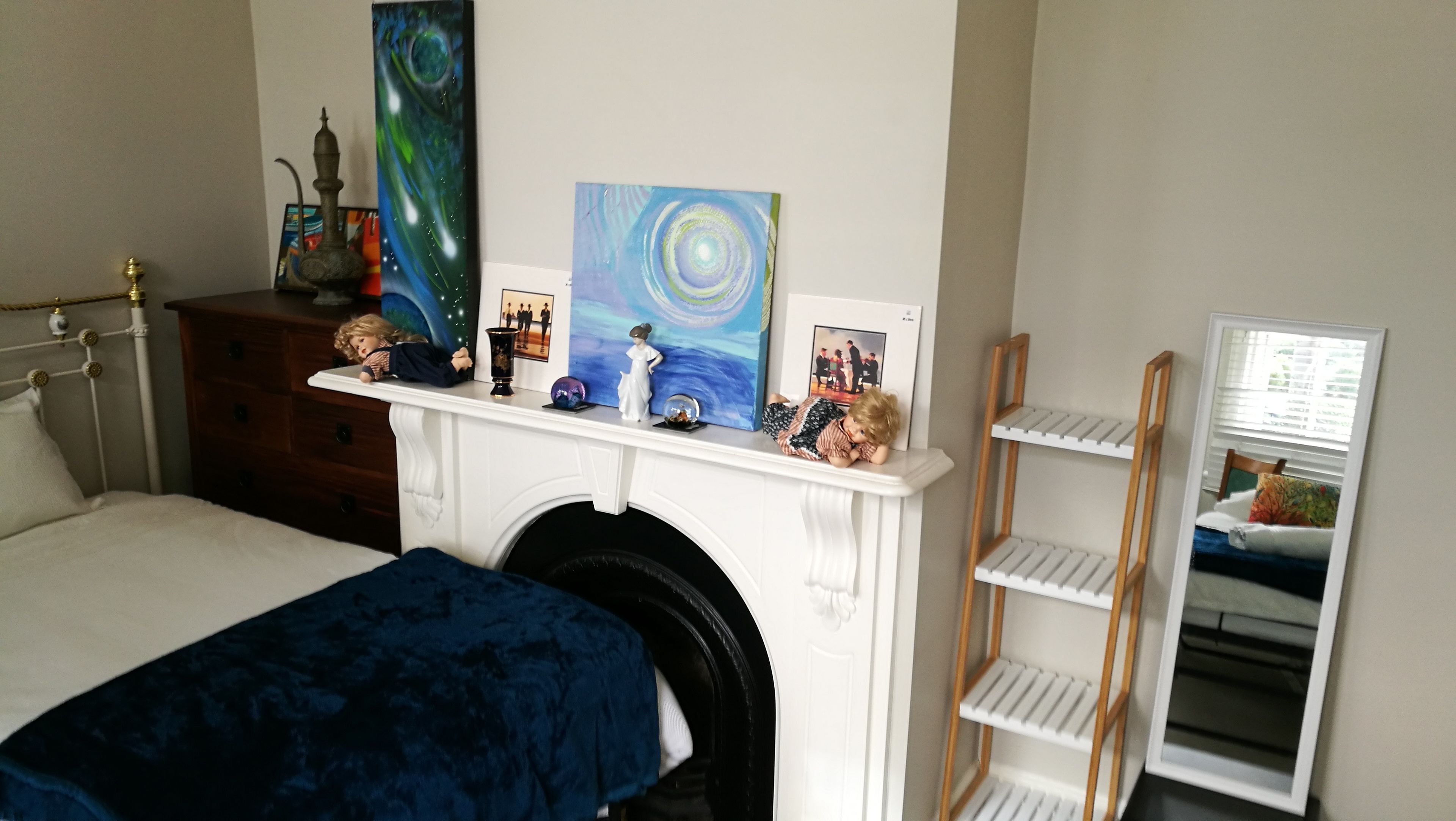 A bedroom with a bed, a dresser, a fireplace mantel with artwork, and a mirror.