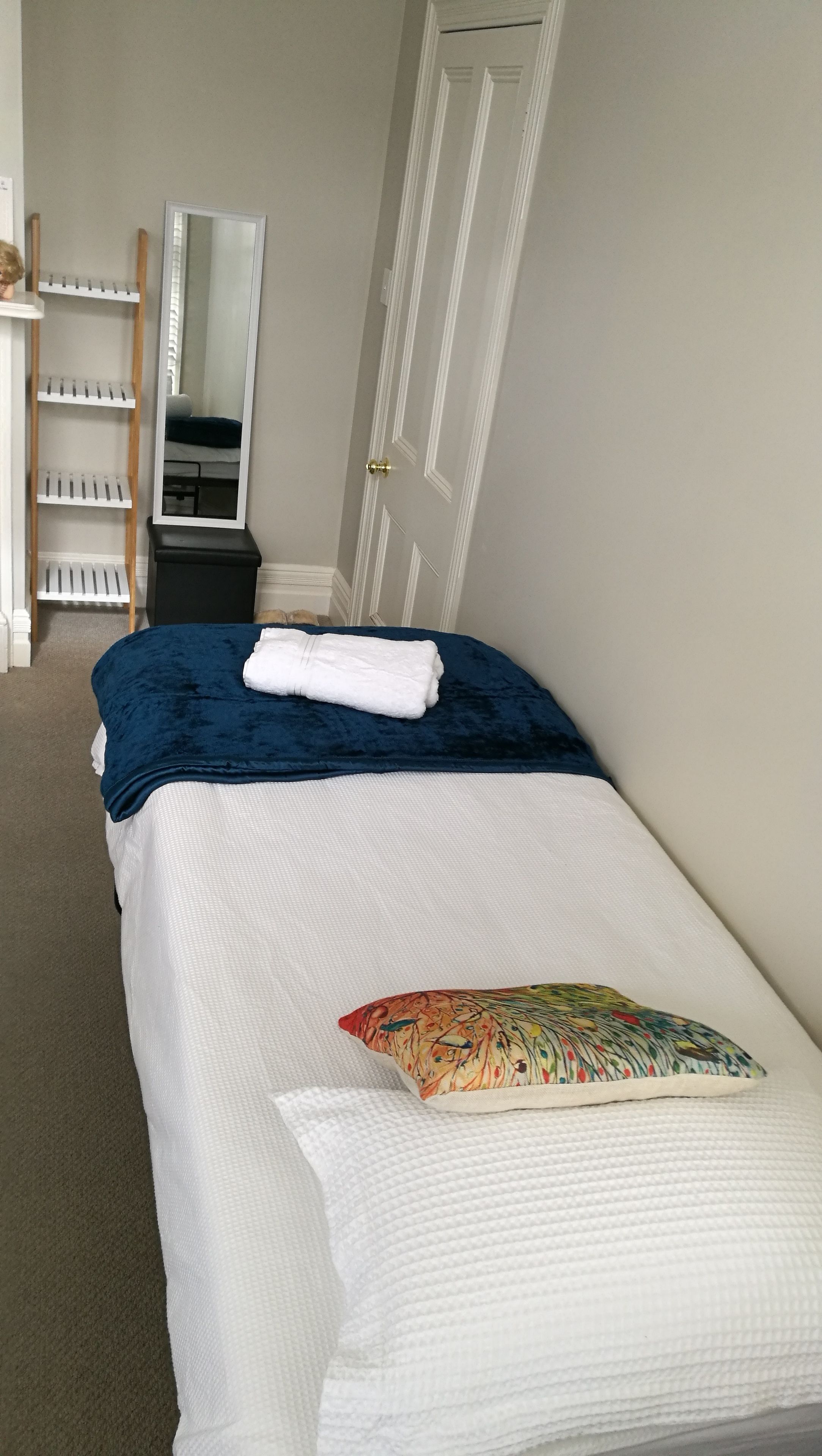 Una cama bien tendida con una manta azul y un cojín colorido, una estantería de madera con libros, un espejo y una puerta en una habitación con paredes de colores claros.