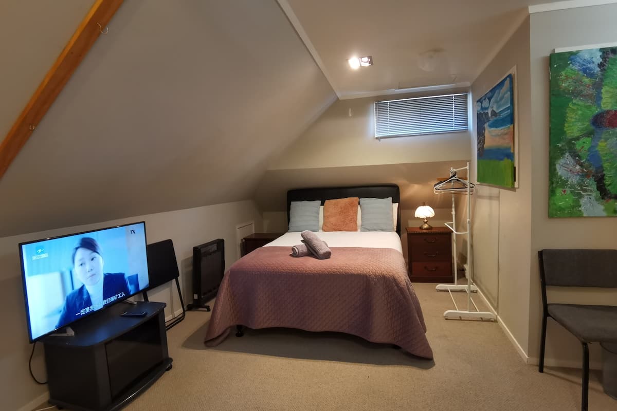 A bedroom with a bed, a TV, a chair, a bedside table, and a painting on the wall.