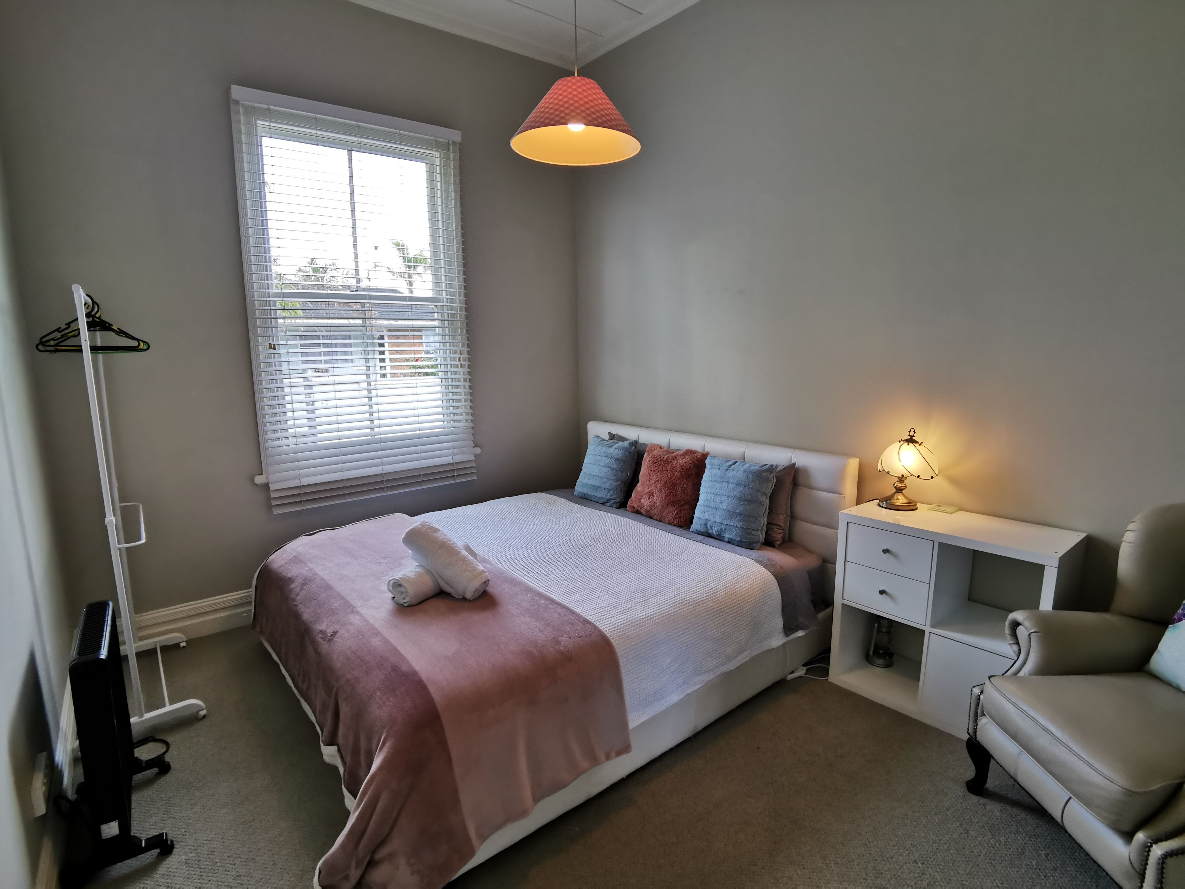 Une chambre à coucher comprenant un lit, une table de chevet, une lampe, une chaise, un radiateur et une fenêtre avec des stores.