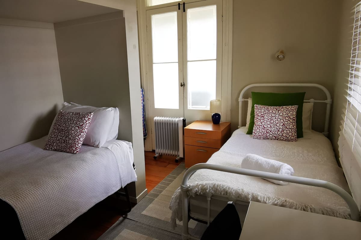 A bedroom with two beds, a radiator, a bedside table with a lamp, and a window with blinds.
