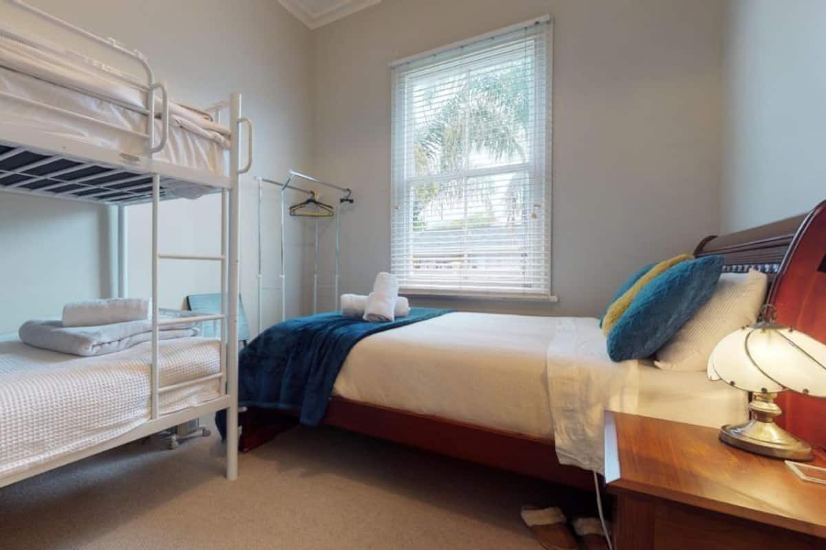 A dormitory room with bunk beds, a bedside table with a lamp, a window with blinds, and a hanging coat rack.