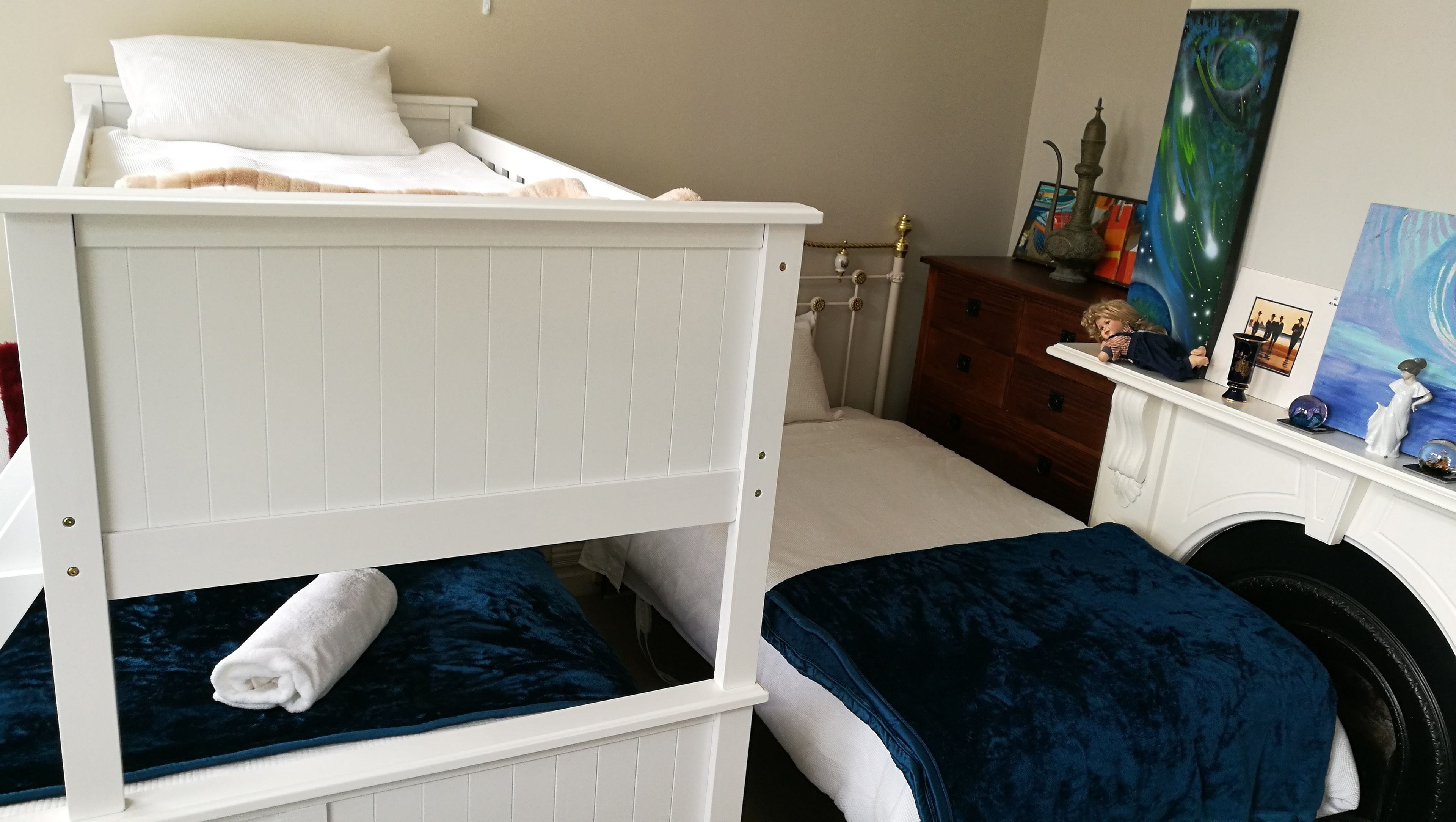 A bunk bed with a white headboard and a lower bunk with a blue blanket. There is a wooden dresser with a mirror and a shelf with decorative items.