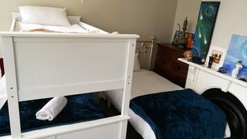 A bunk bed with a white headboard and a lower bunk with a blue blanket. There is a wooden dresser with a mirror and a shelf with decorative items.