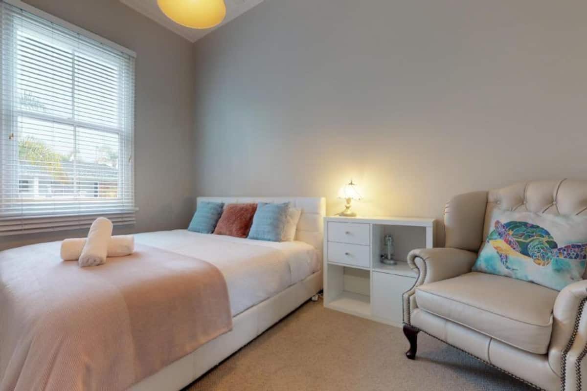 A bedroom with a bed, bedside table, lamp, chair, and window with blinds.