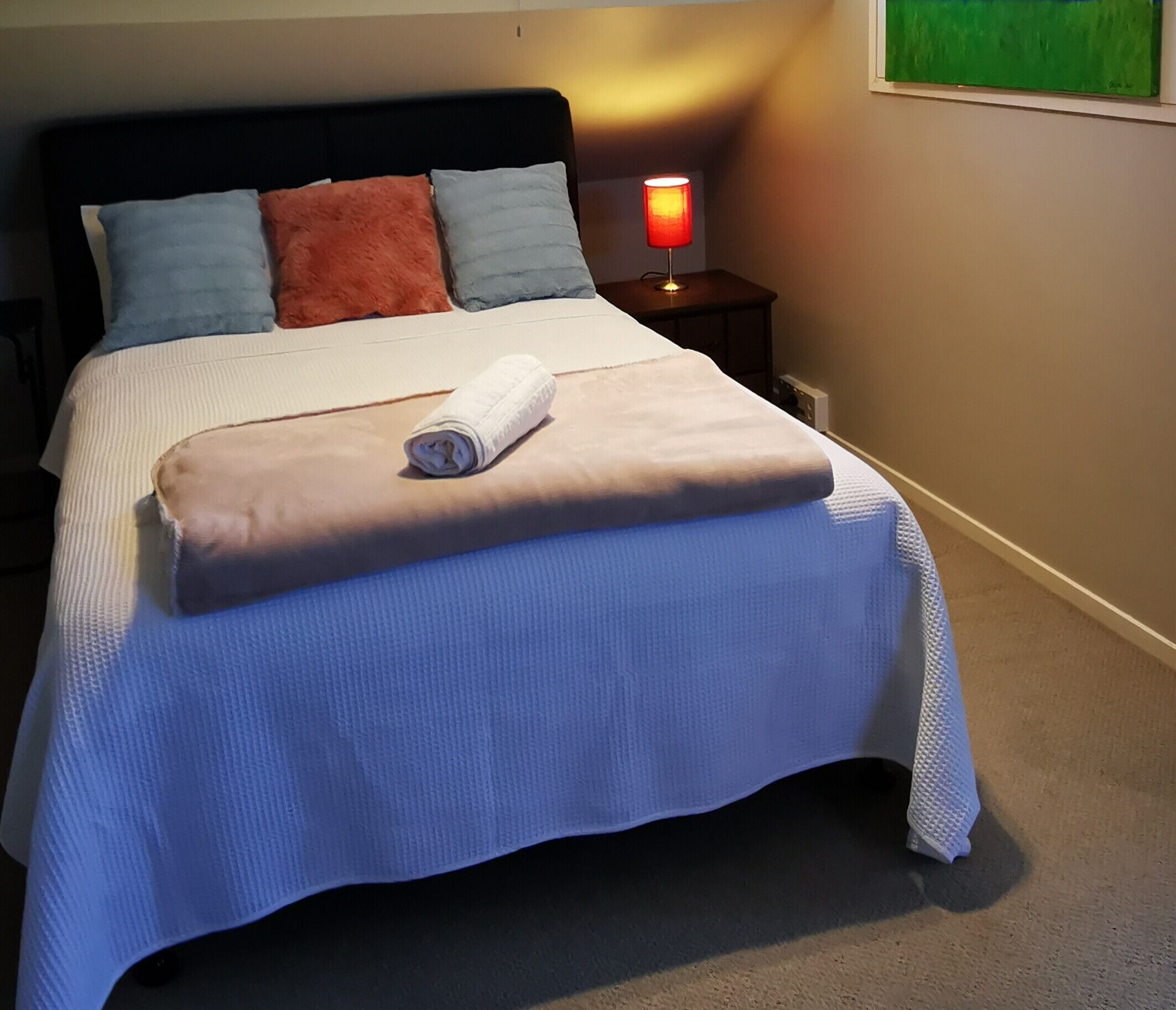 A neatly made bed with a white and blue bedspread, a rolled white towel, and a red lamp on a bedside table.
