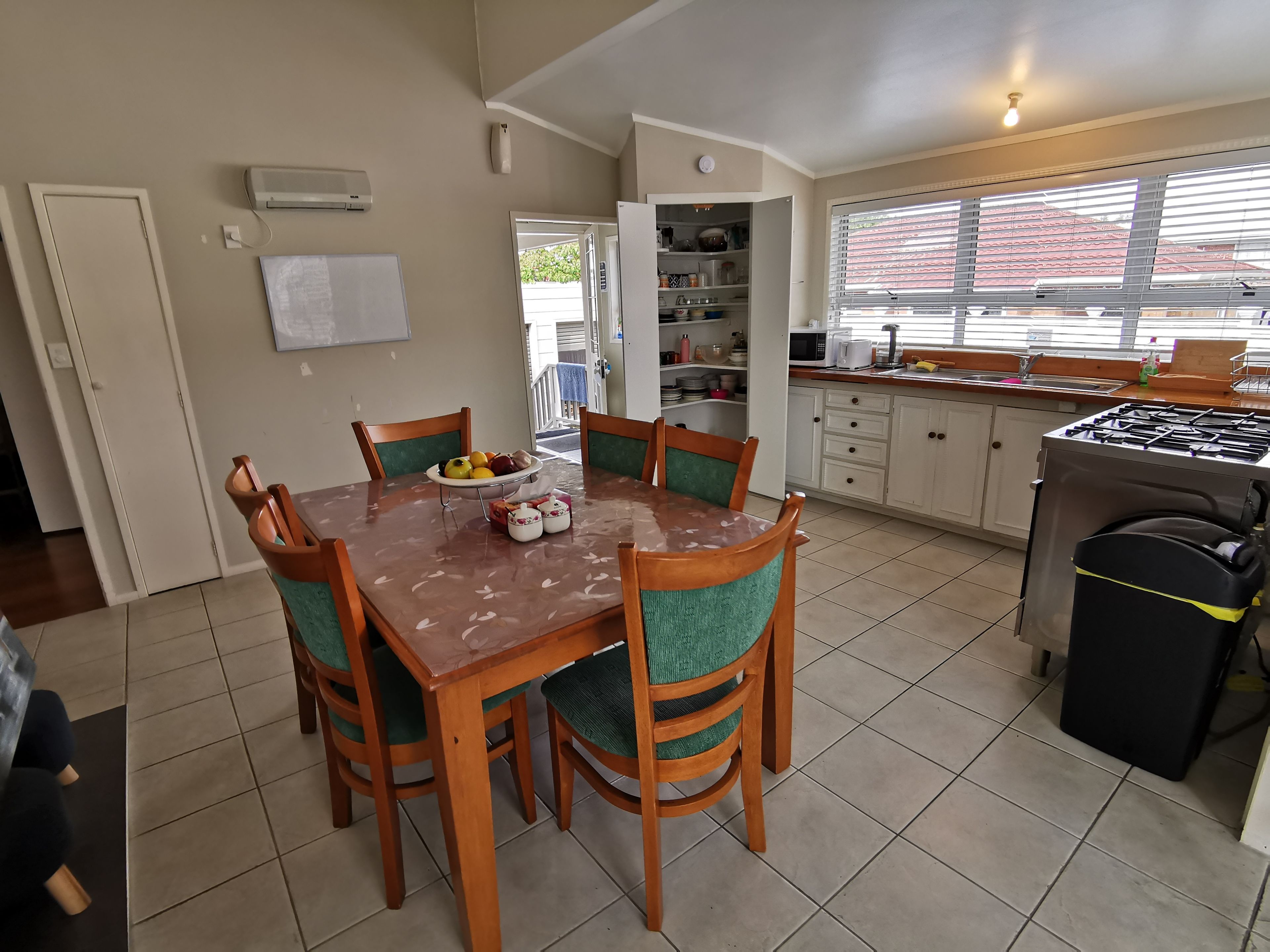 A dining area with a table set for four, surrounded by chairs. A fruit bowl is on the table, and a kitchen with a stove and cabinets is visible in the background.