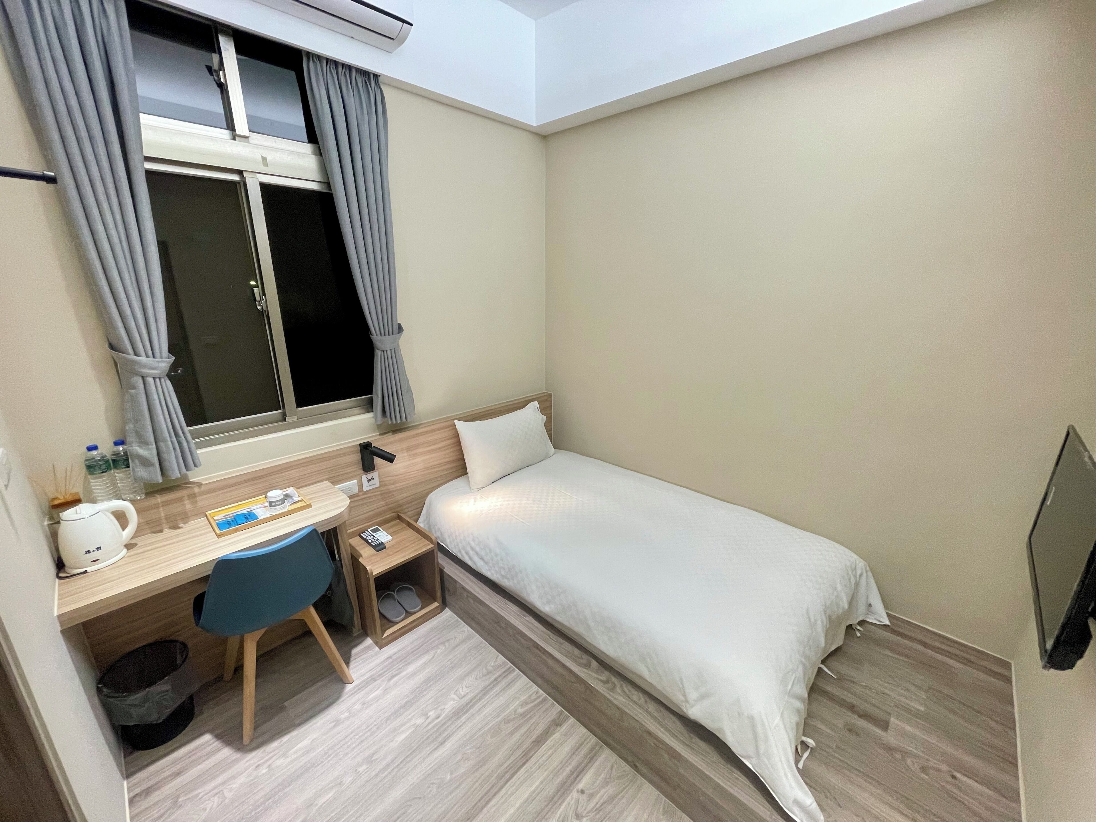 Habitación de hotel compacta con una cama individual, un escritorio con silla, una mesita de noche pequeña, una ventana con cortinas y un equipo de aire acondicionado.