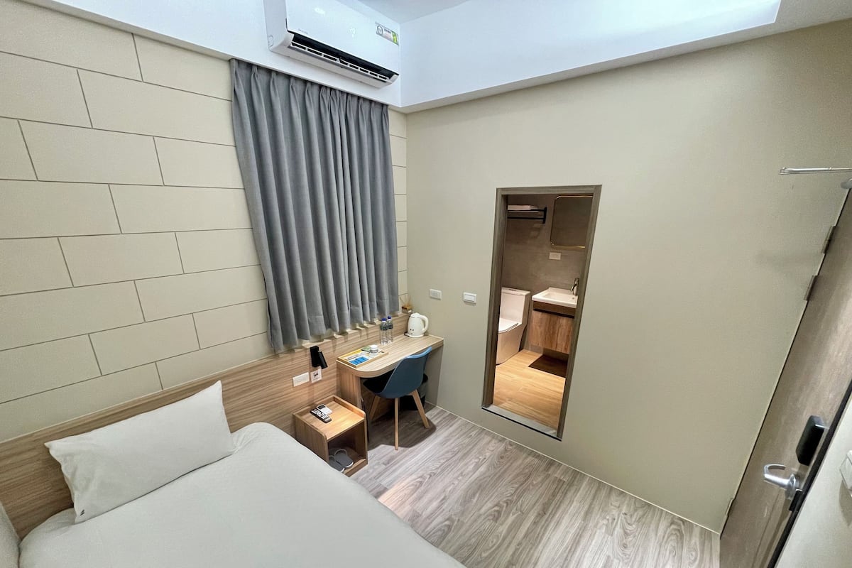 A hotel room with a bed, a desk, a chair, a small table with a remote control, a water bottle, a kettle, and a mirror.