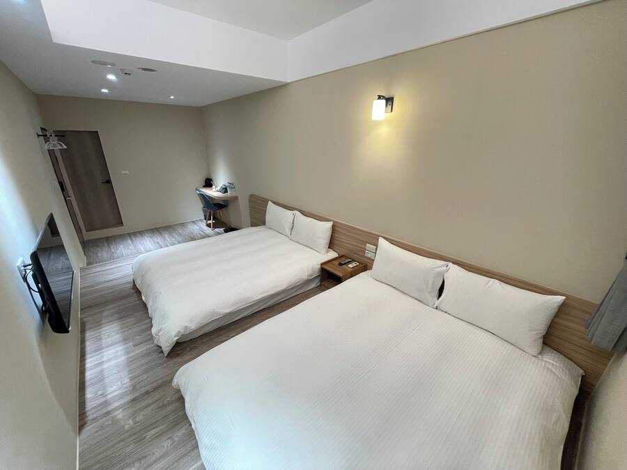 A hotel room with two beds, a desk, a chair, a small table, and a wall-mounted light.