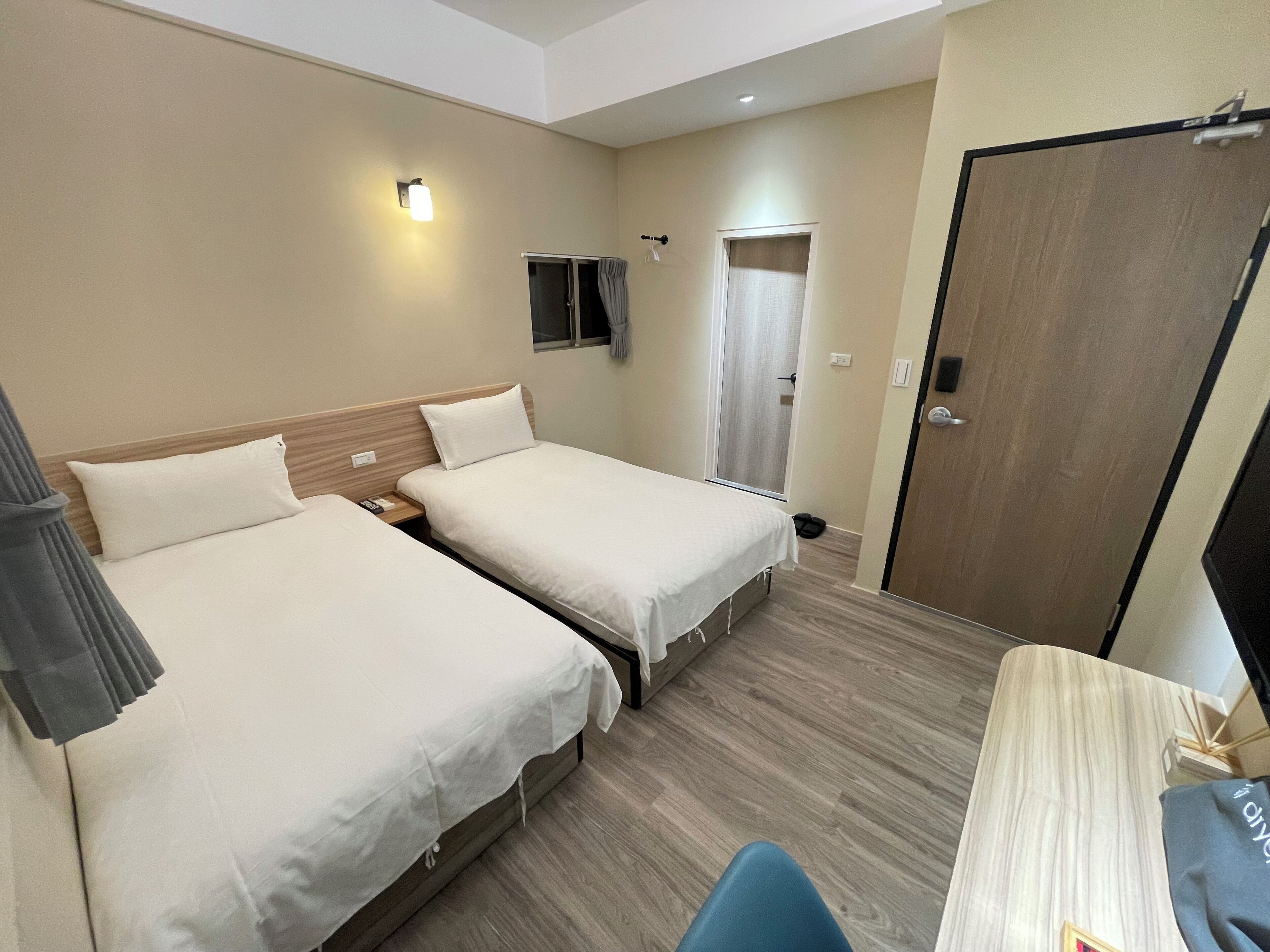 A hotel room with two beds, a desk, a chair, a TV, and a door.