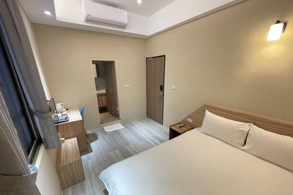 A hotel room with a large bed, a desk with a chair, a wardrobe, and a bathroom visible through an open door.